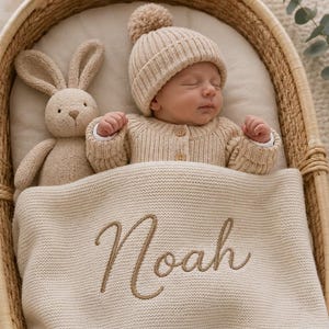 Puede incluir: Un bebé dormido en una cuna tejida, con un gorro y cárdigan de punto beige. Una manta beige bordada con el nombre "Noah" cubre al bebé. Un conejito de peluche descansa junto al bebé, creando una escena de guardería acogedora y de tonos neutros.