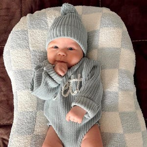 Könnte beinhalten: Ein Baby in einem hellblauen Strick-Outfit, bestehend aus einer Mütze mit Bommel und einem langärmeligen Body. Der Body hat den Namen "ett" auf der Vorderseite gestickt. Das Baby liegt auf einer karierten Decke.