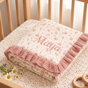 Könnte beinhalten: Eine gefaltete cremefarbene Decke mit einem zarten rosa-grünen Blumenmuster, akzentuiert durch einen gerüschten altrosa Rand. Der Name Maya ist in kursivem rosa Garn in der Mitte des Stoffes eingestickt, der eine strukturierte Oberfläche aufweist.