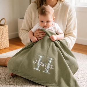 Könnte beinhalten: Eine salbeigrüne Strickdecke für Babys mit einem großen, weiß-grün karierten Buchstaben H, über den der Name Hugo in weißer Schreibschrift gestickt ist. Die Decke hat einen gerippten Rand und eine weiche, strukturierte Strickoberfläche.