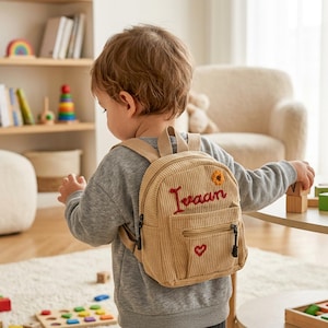 May include: Tan corduroy backpack with the name "Ivaan" embroidered in red. The small backpack features a sunflower and a heart, with a zippered front pocket. Adjustable straps and a top handle are visible.