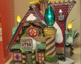 May include: A miniature Christmas village house with a red roof, green trim, and a sign that reads "Brite Lites Bulb Factory". The house has a white snow-covered base and is decorated with lights and greenery.