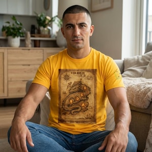 May include: Mustard yellow t-shirt featuring a detailed map design with a dragon and the text "Tolkien". The map is in shades of brown and beige, with the letters "R, G, D, A" overlaid on the dragon. The shirt has a small yellow square on the right sleeve.
