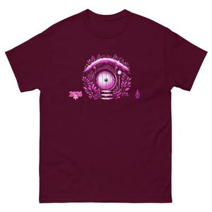 May include: Burgundy t-shirt featuring a pink illustration of a round door set into a hillside, surrounded by foliage. The text "2026" is printed in pink on the left side of the design. A whimsical design for fans of fantasy literature.