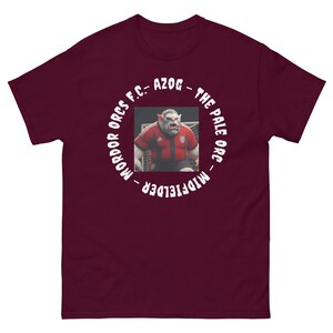 Puede incluir: Camiseta granate con un gráfico de un orco con uniforme de fútbol. El orco lleva una camiseta roja y blanca. El texto alrededor de la imagen dice "MORDOR ORCS F.C. - AZOG - THE PALE ORC - MIDFIELDER."