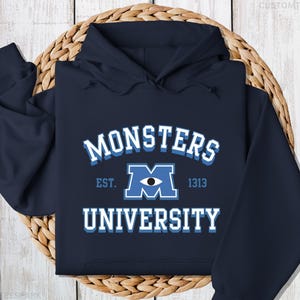 May include: A navy blue hoodie with a white and blue graphic print that says "Monsters University" with a blue "M" logo with a single eye in the center. The text "Est. 1313" is printed below the logo.