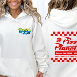 May include: White hoodie featuring a small graphic of three green aliens with blue outfits on the front. The back of the hoodie displays the Pizza Planet logo in red, with the text "FOOD & FUN SPACE PORT" and "SERVING YOUR LOCAL STAR CLUSTER".