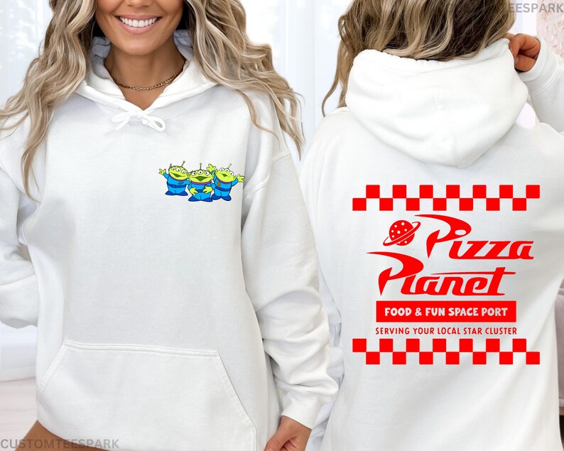 May include: White hoodie featuring a small graphic of three green aliens with blue outfits on the front. The back of the hoodie displays the Pizza Planet logo in red, with the text "FOOD & FUN SPACE PORT" and "SERVING YOUR LOCAL STAR CLUSTER".
