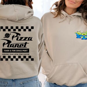 May include: Beige hoodie with a front pocket and drawstring. The back features a black and white checkered pattern above and below the text "Pizza Planet", with "FOOD & FUN SPACE PORT" and "SERVING YOUR LOCAL STAR CLUSTER" below. The front has a small graphic of three green aliens.