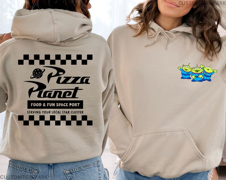 May include: Beige hoodie with a front pocket and drawstring. The back features a black and white checkered pattern above and below the text "Pizza Planet", with "FOOD & FUN SPACE PORT" and "SERVING YOUR LOCAL STAR CLUSTER" below. The front has a small graphic of three green aliens.