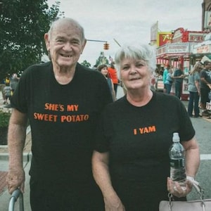 May include: An older couple wearing matching black t-shirts with a sweet potato pun. The man's shirt says "She's my sweet potato" and the woman's shirt says "I yam".