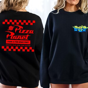 May include: Black sweatshirt featuring the Pizza Planet logo in red on the back, with the text "FOOD & FUN SPACE PORT" and "SERVING YOUR LOCAL STAR CLUSTER". The front displays three green alien characters with blue outfits.
