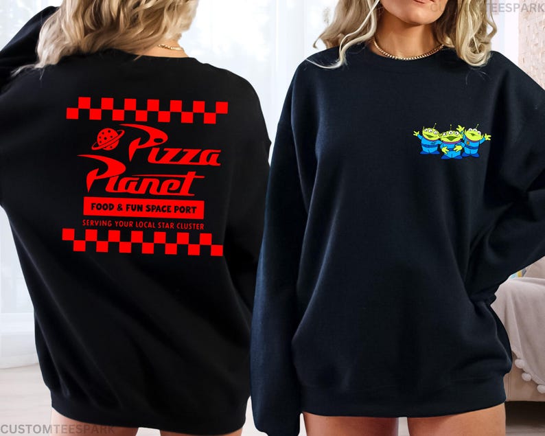 May include: Black sweatshirt featuring the Pizza Planet logo in red on the back, with the text "FOOD & FUN SPACE PORT" and "SERVING YOUR LOCAL STAR CLUSTER". The front displays three green alien characters with blue outfits.