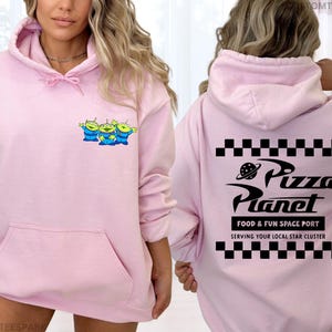 May include: A light pink hoodie featuring a front pocket and a drawstring hood. The front displays a cartoon graphic of three green aliens. The back of the hoodie has the words "Pizza Planet" with a planet graphic and the text "FOOD & FUN SPACE PORT".
