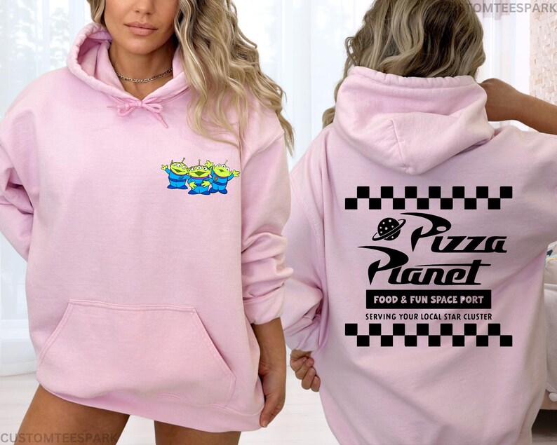 May include: A light pink hoodie featuring a front pocket and a drawstring hood. The front displays a cartoon graphic of three green aliens. The back of the hoodie has the words "Pizza Planet" with a planet graphic and the text "FOOD & FUN SPACE PORT".