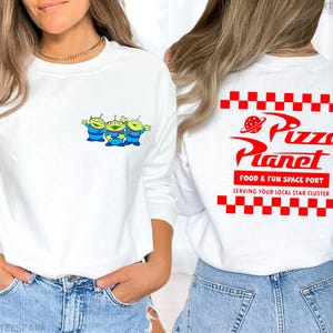 May include: White long-sleeve sweatshirt featuring a small graphic of three green aliens in blue suits on the front. The back of the shirt displays the Pizza Planet logo in red, with the text "FOOD & FUN SPACE PORT" and "SERVING YOUR LOCAL STAR CLUSTER".