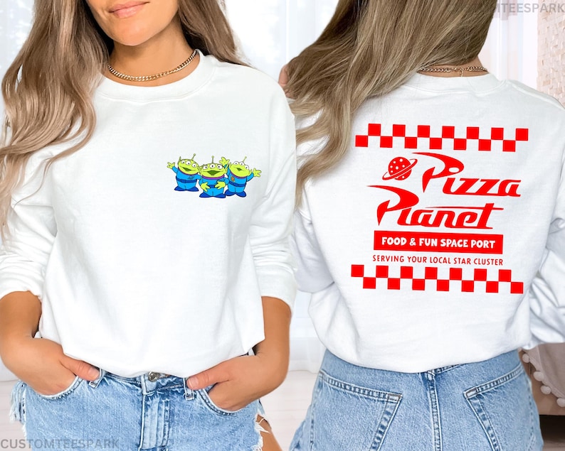 May include: White long-sleeve sweatshirt featuring a small graphic of three green aliens in blue suits on the front. The back of the shirt displays the Pizza Planet logo in red, with the text "FOOD & FUN SPACE PORT" and "SERVING YOUR LOCAL STAR CLUSTER".