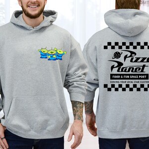 May include: A light gray hoodie featuring a front pocket and a drawstring hood. The front displays three cartoon green aliens in blue suits. The back of the hoodie has the text "Pizza Planet" with a planet graphic and checkerboard patterns.