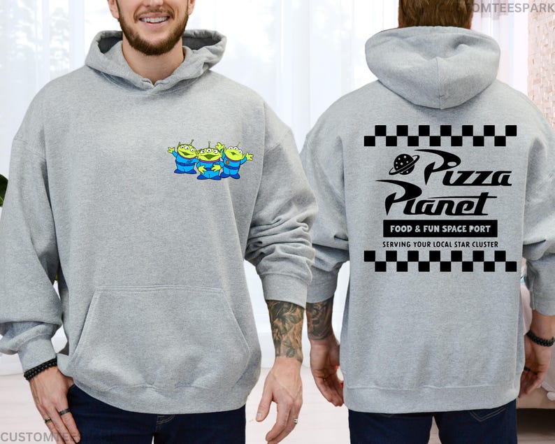 May include: A light gray hoodie featuring a front pocket and a drawstring hood. The front displays three cartoon green aliens in blue suits. The back of the hoodie has the text "Pizza Planet" with a planet graphic and checkerboard patterns.