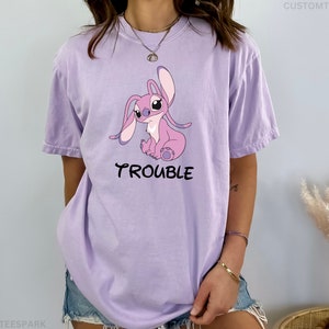 May include: A lavender t-shirt with a cartoon image of Stitch, a pink alien character with big ears, sitting down. The text "Trouble" is printed below the image.
