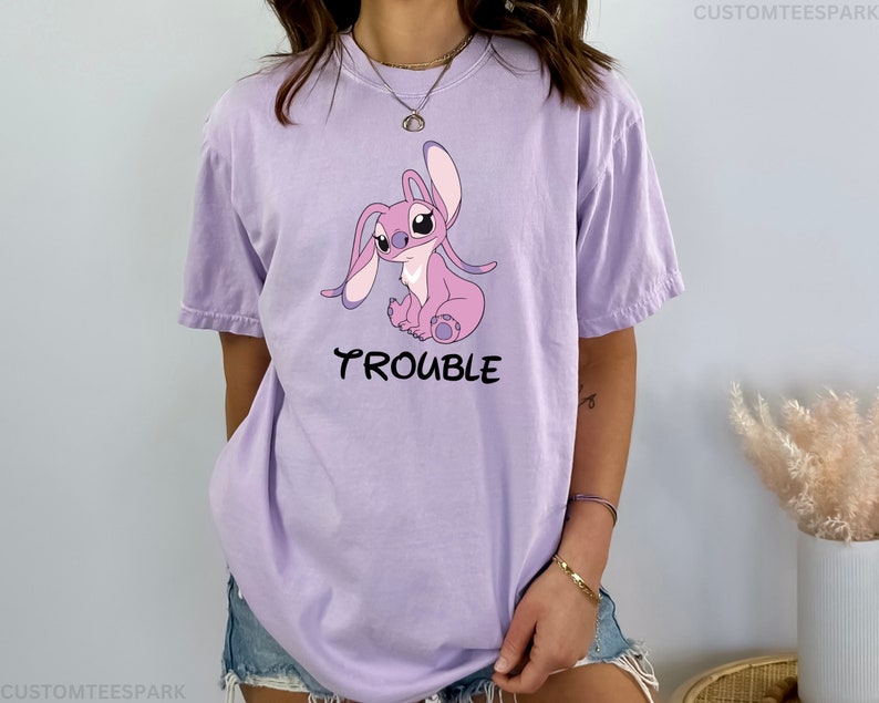 May include: A lavender t-shirt with a cartoon image of Stitch, a pink alien character with big ears, sitting down. The text "Trouble" is printed below the image.