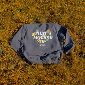 May include: A gray sweatshirt with the text "THAT'S MOCKED UP" and various colorful graphic designs. The sweatshirt is lying on a bed of yellow flowers, creating a vibrant contrast. The words "Premium Mock Ups" are also visible.