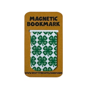 Puede incluir: Un marcapáginas magnético con un patrón repetido de tréboles verdes de cuatro hojas sobre un fondo blanco. El marcapáginas está adherido a un respaldo de cartón marrón con las palabras "MAGNETIC BOOKMARK" impresas en negro. La dirección del sitio web www.zestypineappleshop.com está impresa en la parte inferior.