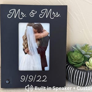 May include: Black picture frame with the words "Mr. & Mrs." and the date "9/9/22" in white script. The frame holds a wedding photo of a couple embracing. A small speaker is visible on the frame. A decorative plant sits to the right.