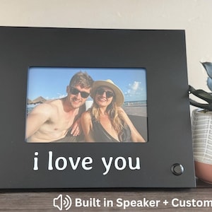 May include: Black photo frame with a picture of a couple at the beach. The words "i love you" are printed in white at the bottom. A small speaker button is on the right side. The text "Built in Speaker + Custom Text" is at the bottom.