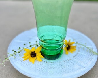 May include: A green glass shot glass with a white and gold rim sits on a white cake stand with a gold floral design. Two yellow flowers are on the stand.
