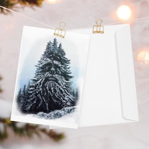 Könnte beinhalten: Eine Grußkarte mit einer verschneiten Waldszene, befestigt mit einer goldenen Büroklammer an einer Schnur. Ein weißer Umschlag befindet sich neben der Karte. Der Hintergrund zeigt weiche, verschwommene Lichter und einen verschneiten Zweig.