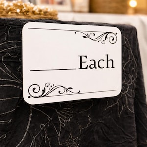 May include: A white rectangular sign with rounded corners, displaying the word "Each" in a bold, black font. The sign has decorative black flourishes at the top and bottom. The sign is placed on a dark, textured surface.