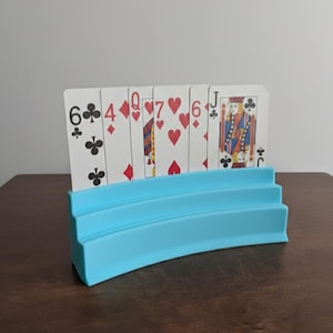 May include: A light blue plastic playing card holder with a curved design, holding a hand of playing cards. The cards show various suits and numbers, including a Jack and a 6 of clubs.