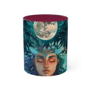 May include: A maroon ceramic mug with a colorful illustration of a woman's face surrounded by foliage and a full moon. The woman's eyes are closed and she has a serene expression.