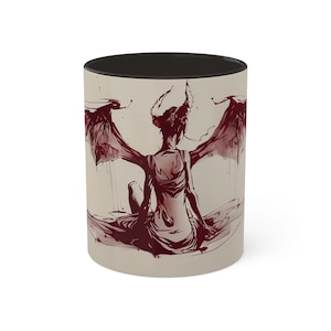 Puede incluir: Una taza de cerámica blanca con un borde y un asa negros. La taza presenta una ilustración roja y blanca de una mujer con alas y cuernos, sentada sobre una superficie roja.