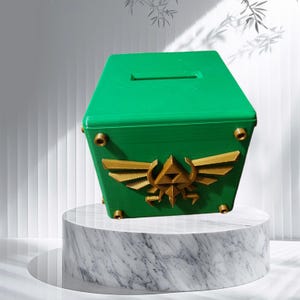 May include: A green treasure chest with a gold Triforce symbol on the front. The chest has a slot on the top for inserting coins.