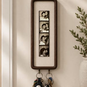 May include: A vertical rectangular dark wood wall-mounted key holder featuring a central cream-colored fabric panel. The panel displays a vertical strip of four black and white photo booth images. Two metal hooks at the bottom hold sets of keys.