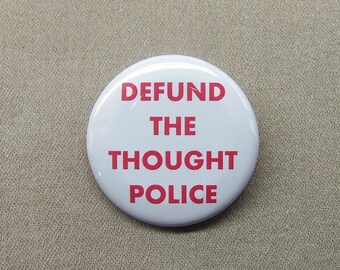 May include: A white button with the text "DEFUND THE THOUGHT POLICE" in red. The button is round and has a glossy finish. The background is a neutral tan color.