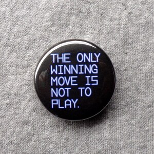May include: A round, black button with the text "THE ONLY WINNING MOVE IS NOT TO PLAY" in white. The button is centered on a gray, textured fabric background.