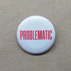 May include: A white, round button with the word "PROBLEMAtIC" printed in red, centered on the face. The button is set against a neutral, tan background.