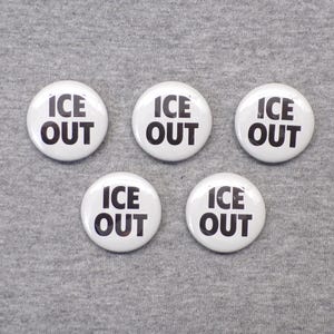 May include: Five white round buttons with the words "ICE OUT" in bold black letters. The buttons are arranged on a gray surface. The buttons are a simple, clean design.