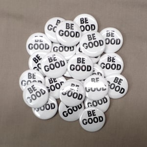 May include: A collection of white round buttons with the words "BE GOOD" printed in black. The buttons are scattered on a neutral brown surface. The text is centered on each button.