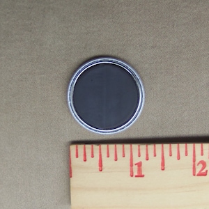 May include: A round, silver-colored button with a black, magnetic back. The button is about 1 inch in diameter.