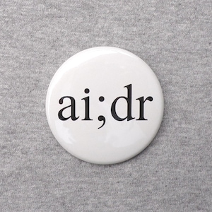 May include: A white, round button with the text "ai;dr" in black, centered on the button. The button is set against a gray, textured fabric background.