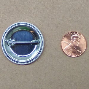 May include: The back of a round button with a silver metal backing and a pin on the back. The button is about the size of a US penny.