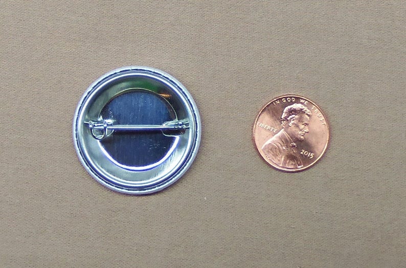 May include: The back of a round button with a silver metal backing and a pin on the back. The button is about the size of a US penny.