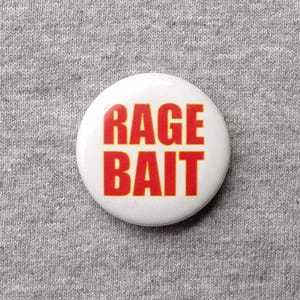 May include: A white button with the words "RAGE BAIT" in bold red letters. The button is centered on a gray, textured fabric background.