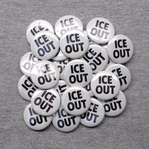 May include: A collection of white round buttons with the words "ICE OUT" printed in bold black letters. The buttons are scattered on a gray fabric surface, creating a visually appealing arrangement.