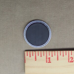 May include: A round, silver-colored button with a black, magnetic back. The button is about 1 inch in diameter.