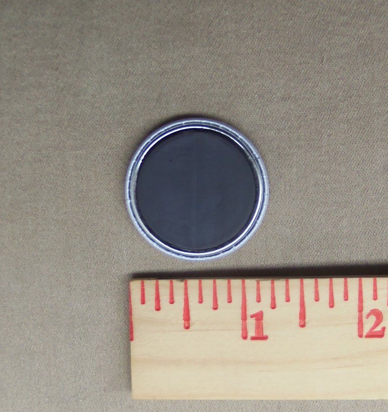 May include: A round, silver-colored button with a black, magnetic back. The button is about 1 inch in diameter.